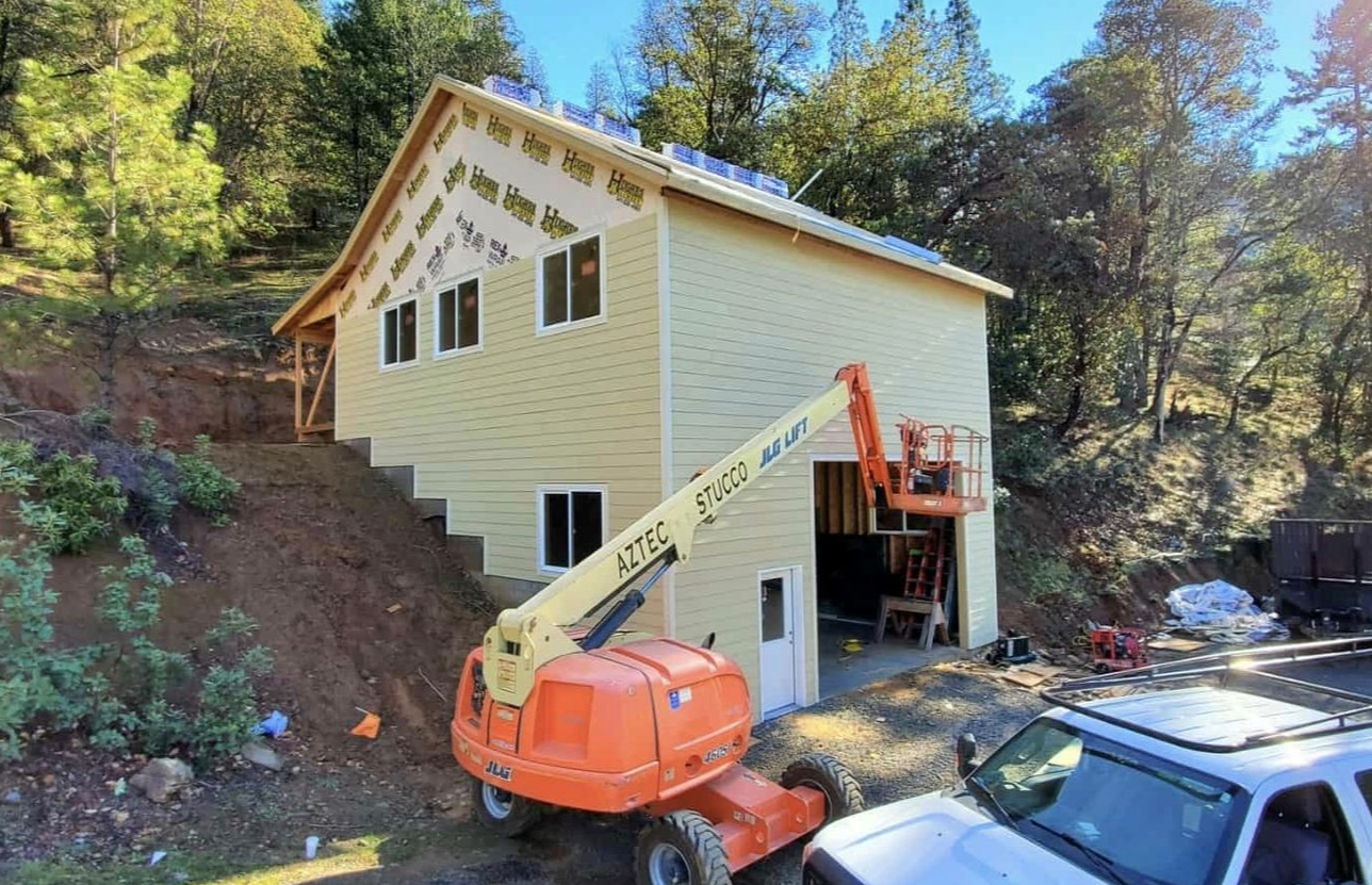 A two-story building under construction. Yellow siding, a worker in a lift, and a white truck in front.