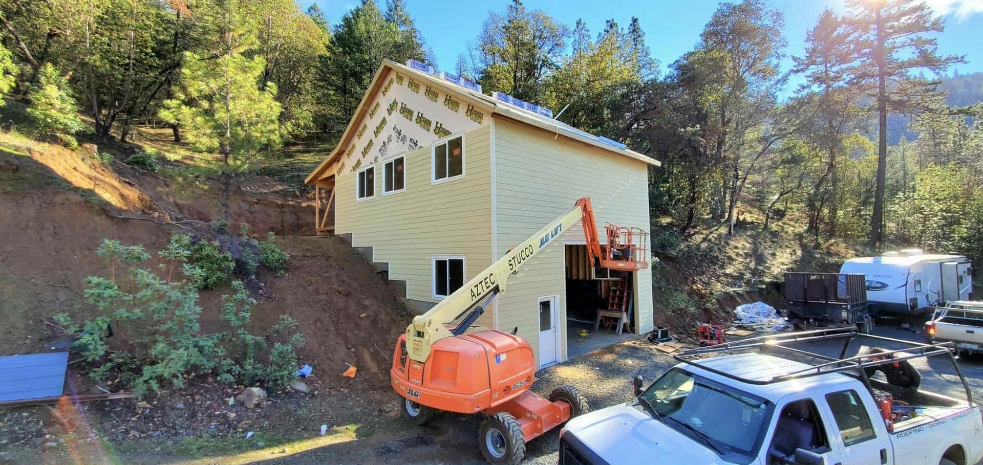 Construction site with building, lift, trucks, and trees on a sunny day.