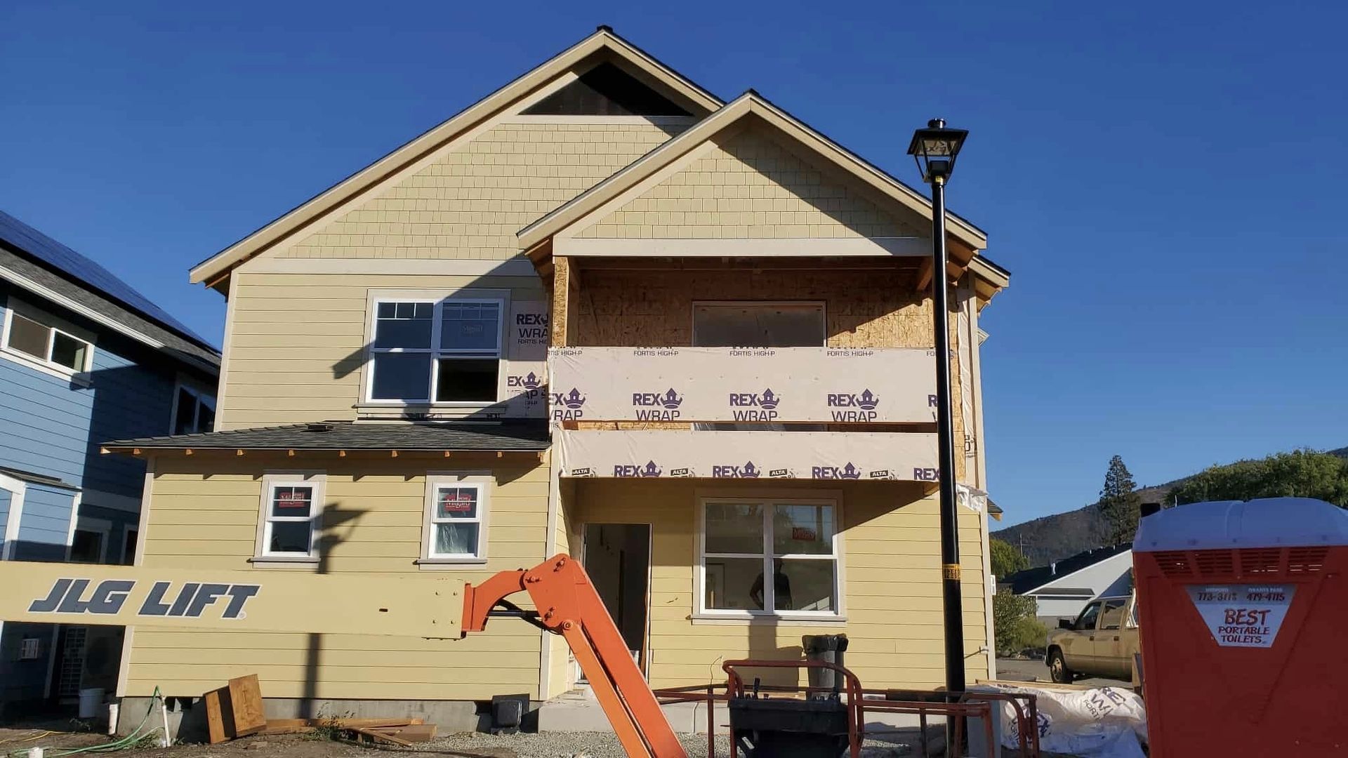 Two-story house under construction, beige siding, balcony, windows, clear sky, construction equipment.