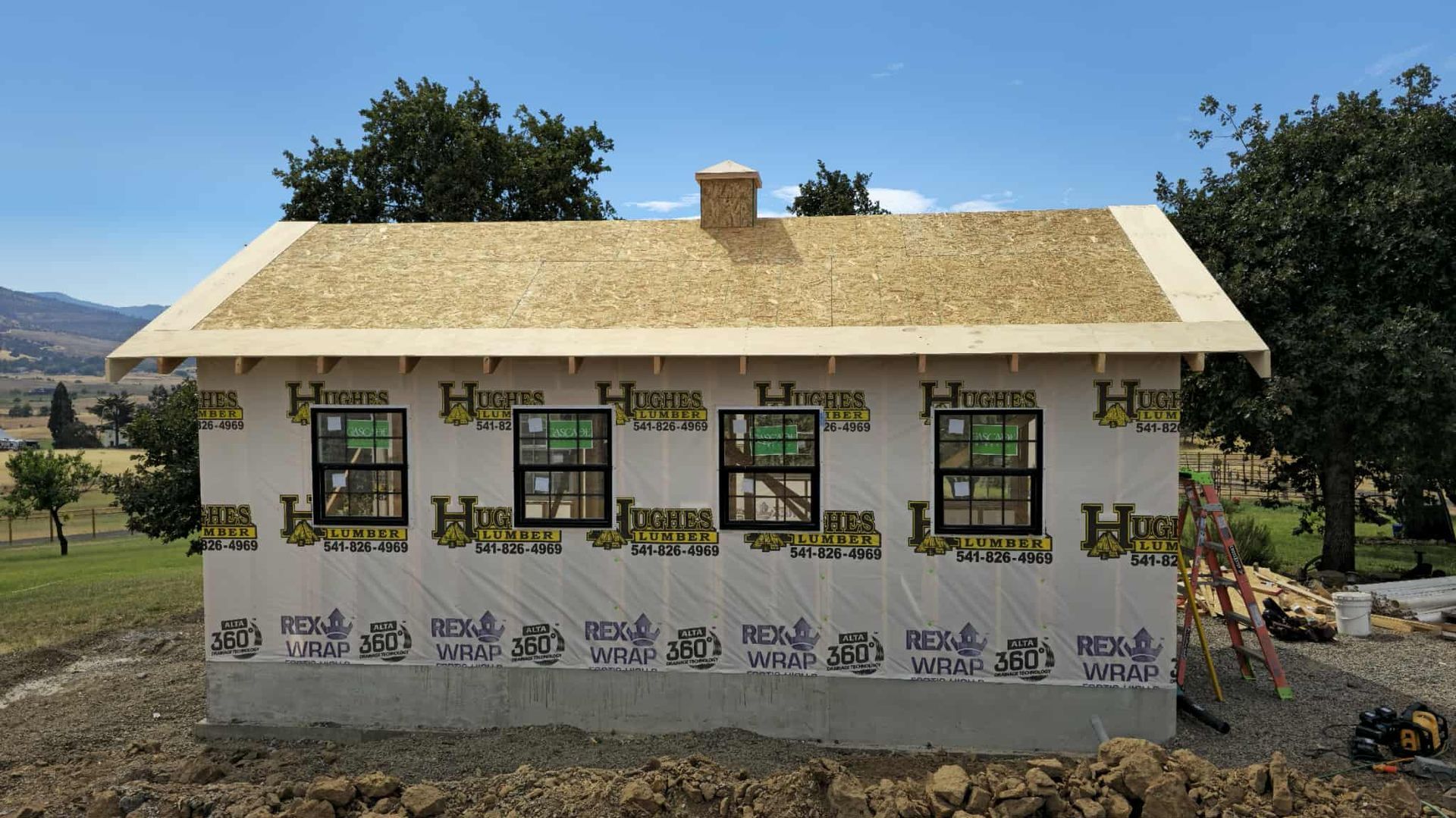 A small house under construction with windows, a roof, and a chimney, set in a rural landscape.
