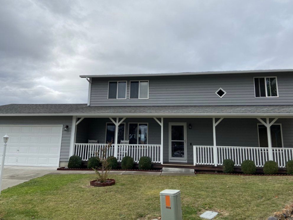 Gray two-story house with white trim and a porch, on a cloudy day, with lawn and shrubs.