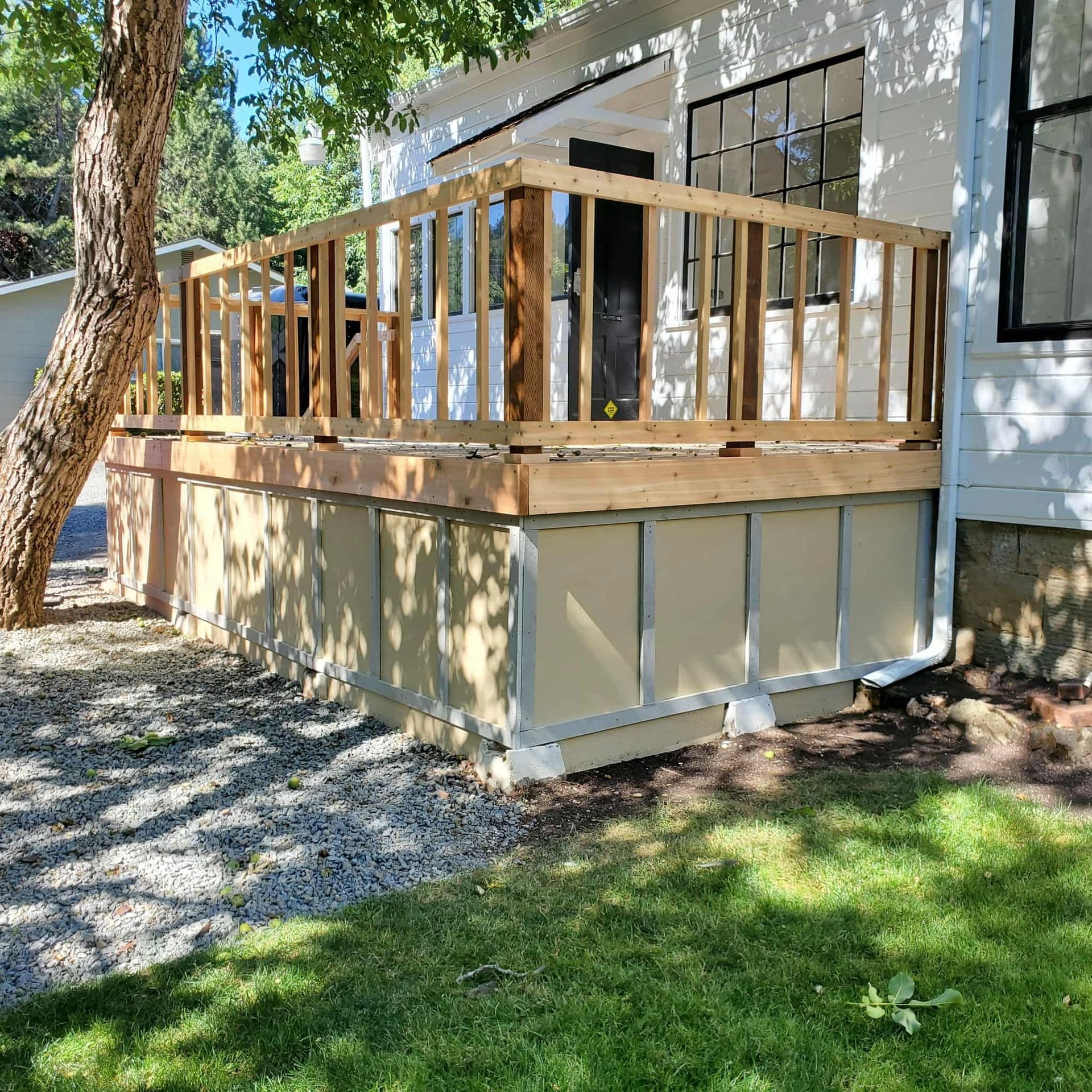 A wooden deck with railings is attached to a white house, over a skirted base, surrounded by grass and gravel.