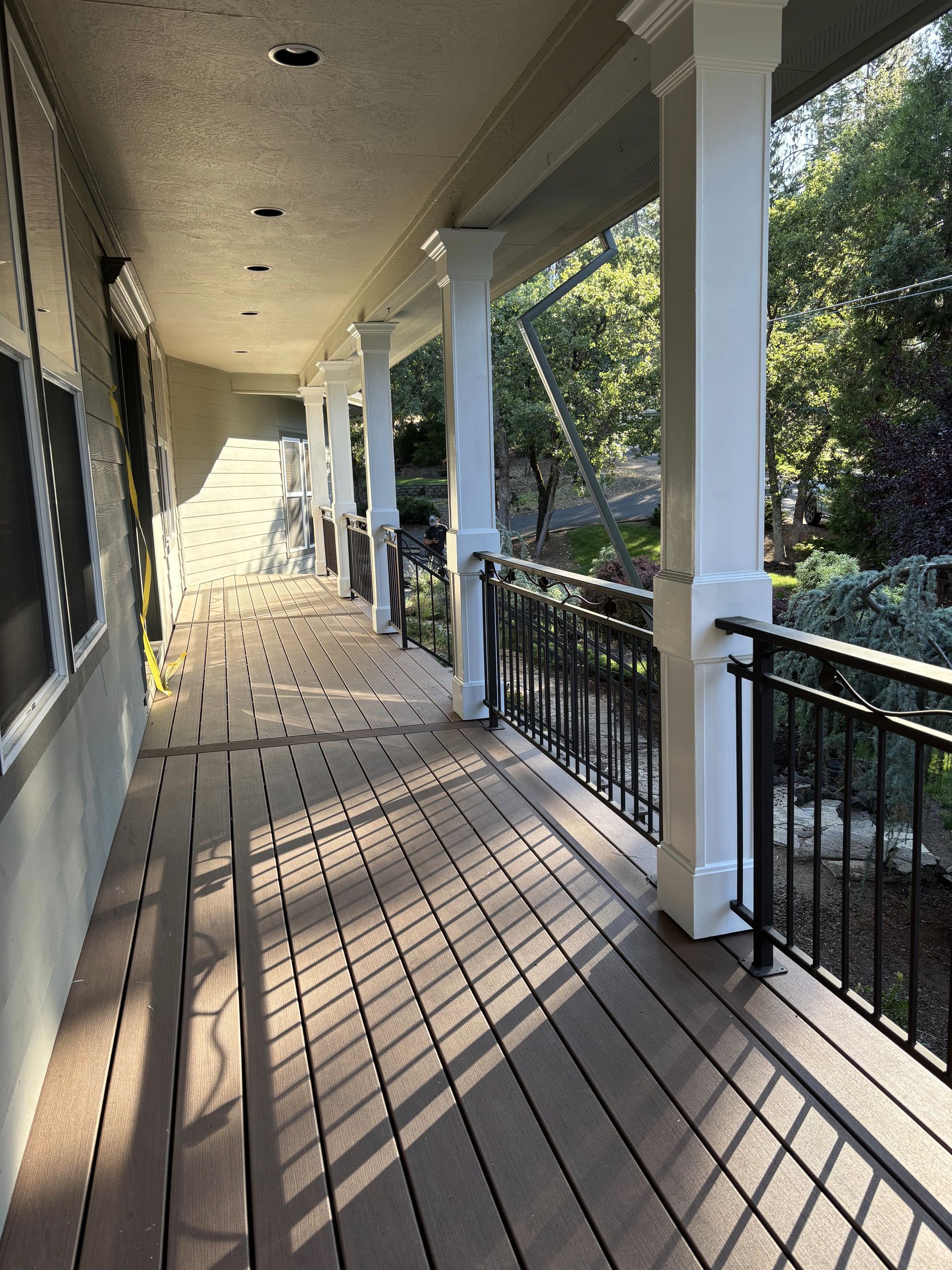A long covered porch with brown decking, white pillars, and black railings. Sunlight casts shadows.