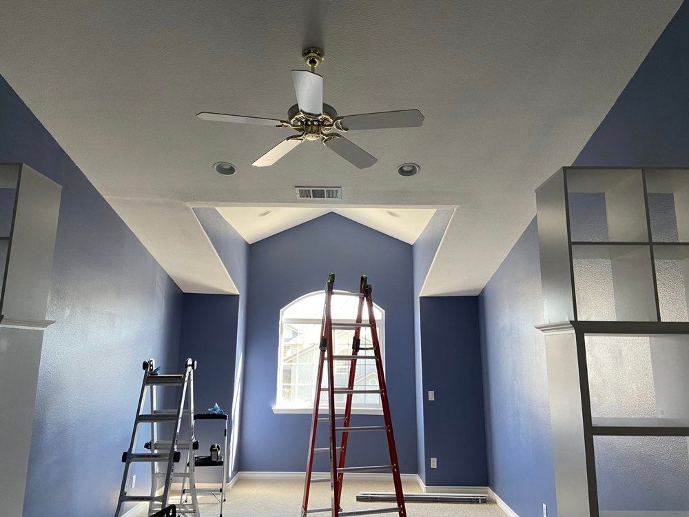 Room with blue walls, white ceiling, ladder near window, fan.