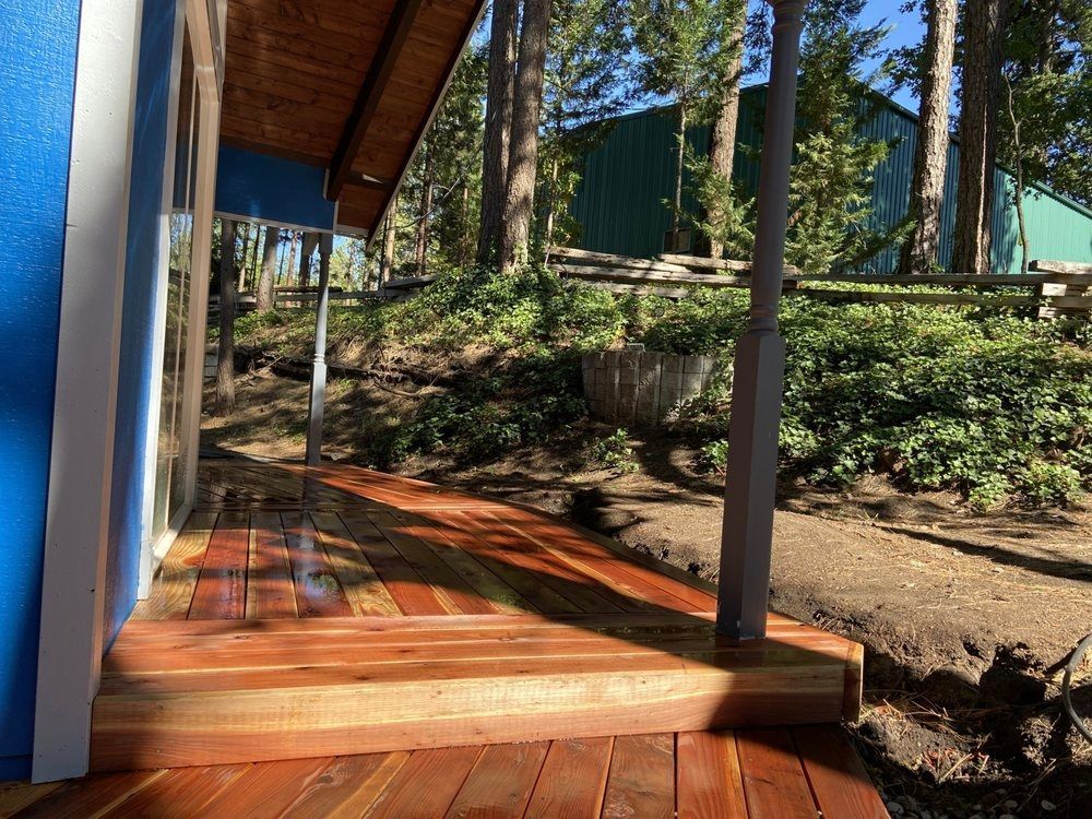 Wooden deck in front of a blue building, with a natural hillside and trees in the background.