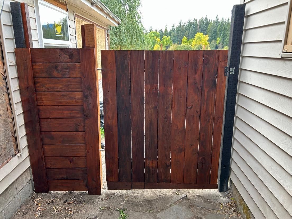 Wooden gate in a brown finish. It is open between two dark posts, next to a siding building.