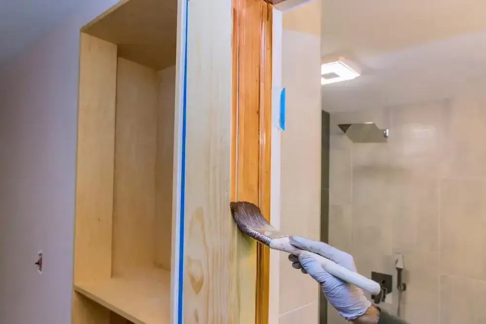 Person staining a wooden cabinet with a brush, wearing gloves. Bathroom setting.