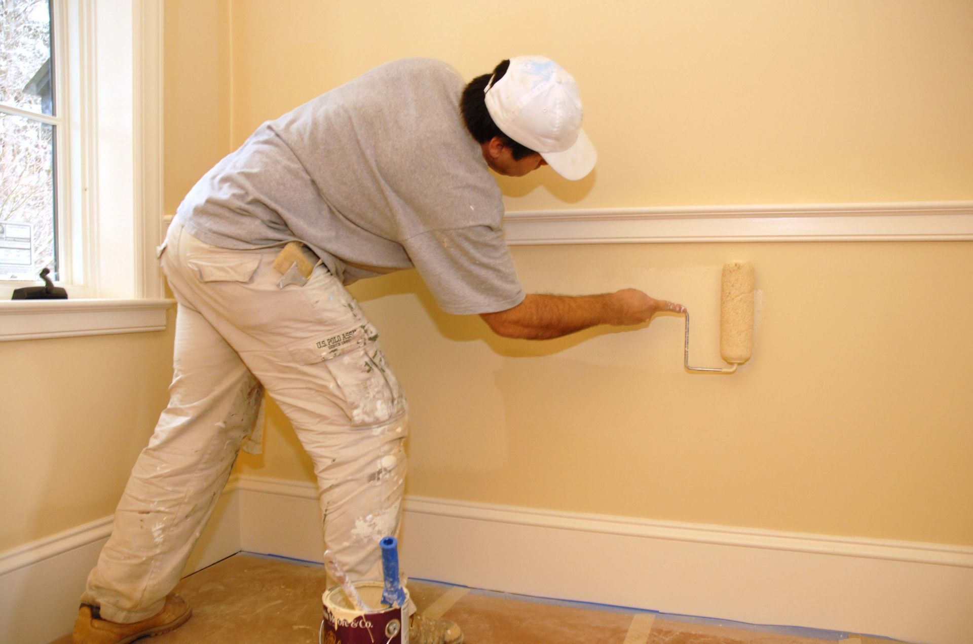 Person paints a yellow interior wall with a roller. White trim and window are visible.