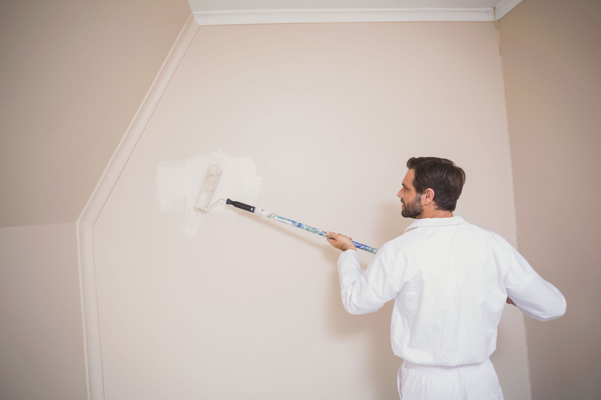 Man in white coveralls paints a wall with a roller. Beige walls and white trim in a room.