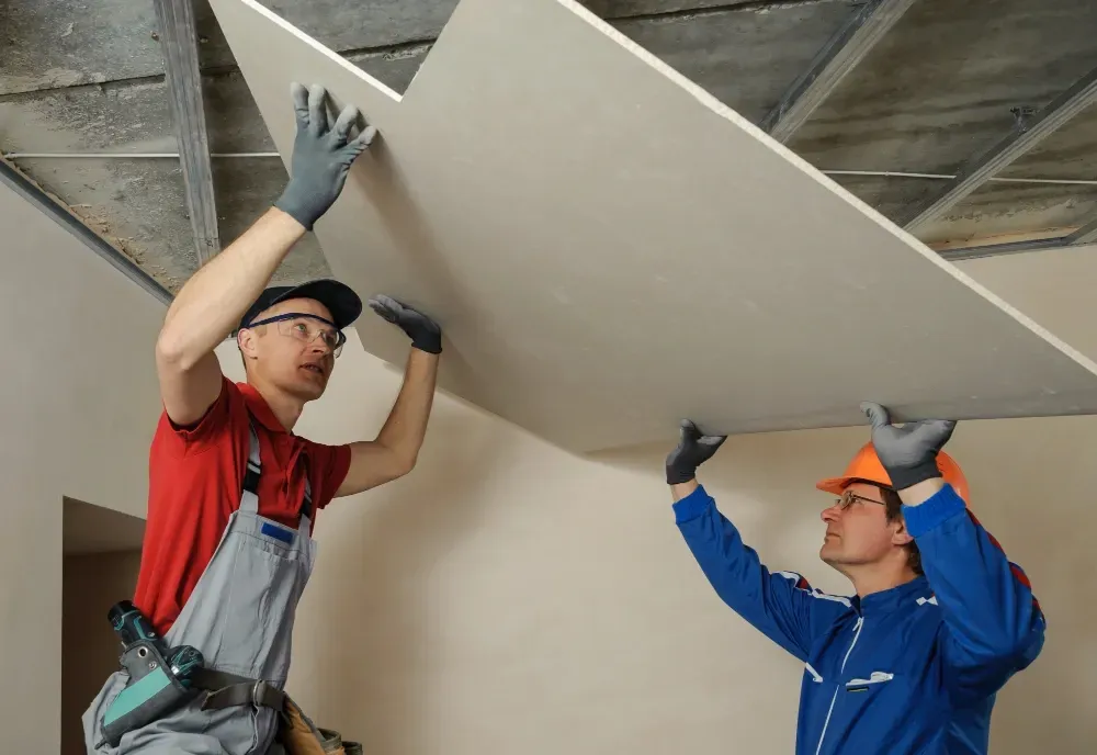 Two construction workers installing a drywall ceiling. One holds the panel while the other guides it.