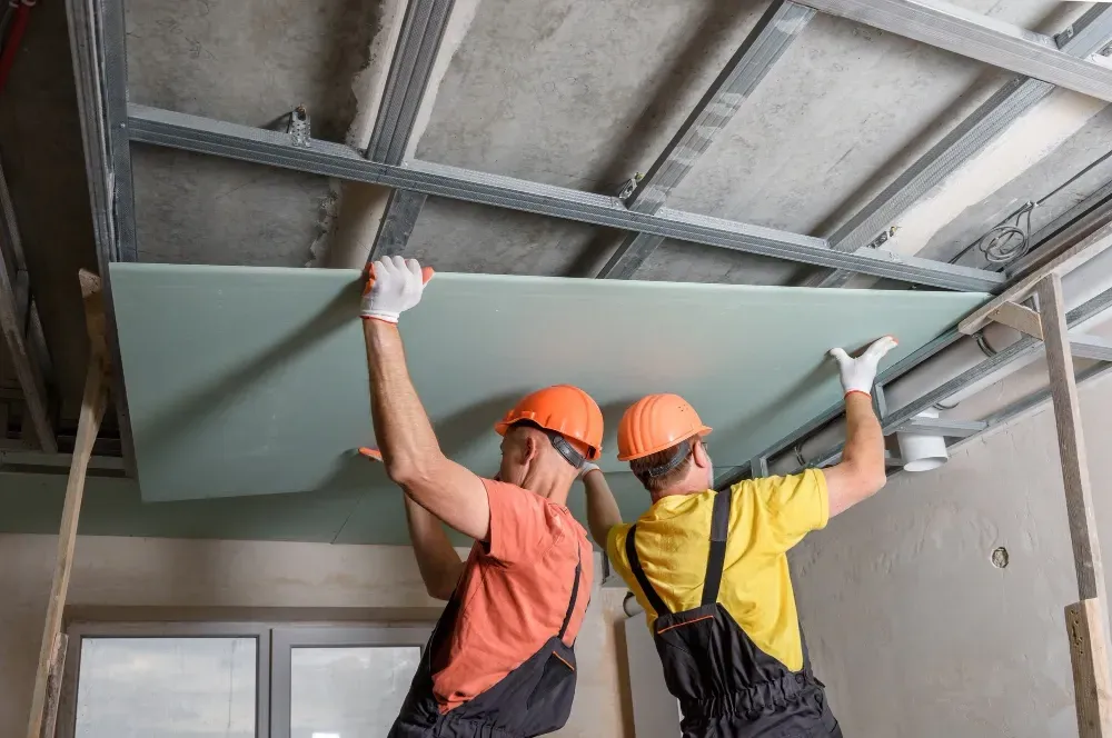 Two construction workers installing drywall on a ceiling, wearing hard hats and gloves.