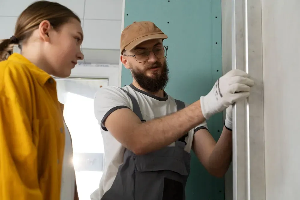 A construction worker in overalls and cap shows a woman drywall installation.