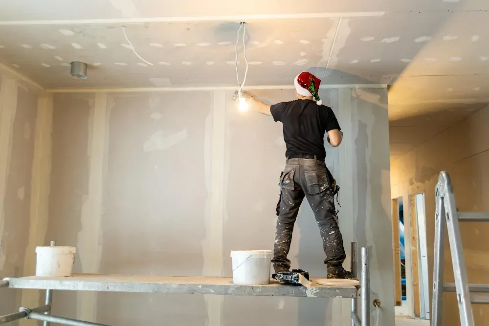 Construction worker in Santa hat, on scaffolding, working on drywall in a room.