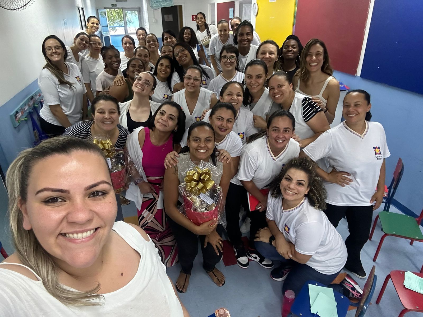 Grupo de mulheres sorrindo em uma sala de aula; muitas vestindo camisas brancas, algumas segurando presentes.