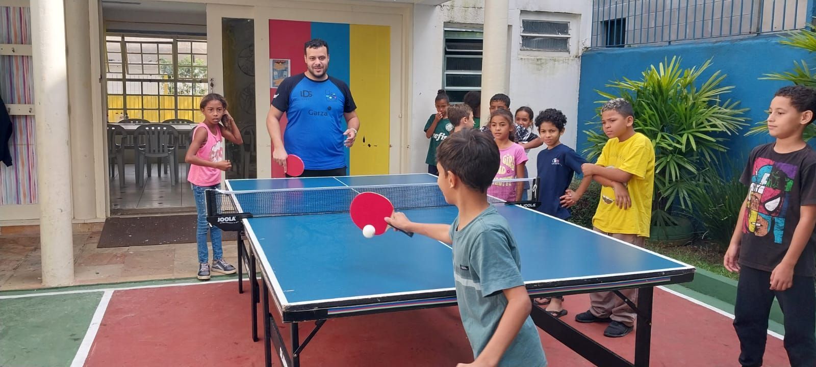 A group of children watching a table tennis match between a man and a boy. The game is outdoors.