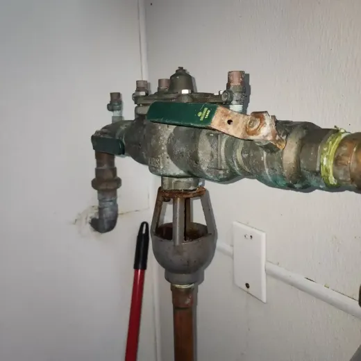 A corroded backflow preventer valve with copper and galvanized pipes attached to a white wall.