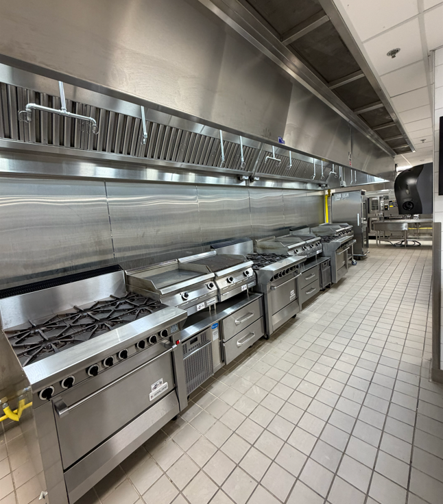 Commercial kitchen with stainless steel stoves, ovens, and ventilation hood along a tiled wall.