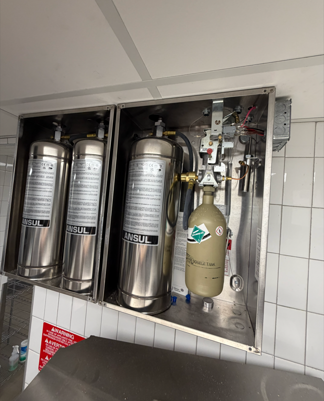 Open wall cabinet with metal cylinders and a beige gas tank, showing valves and tubing inside a tiled room