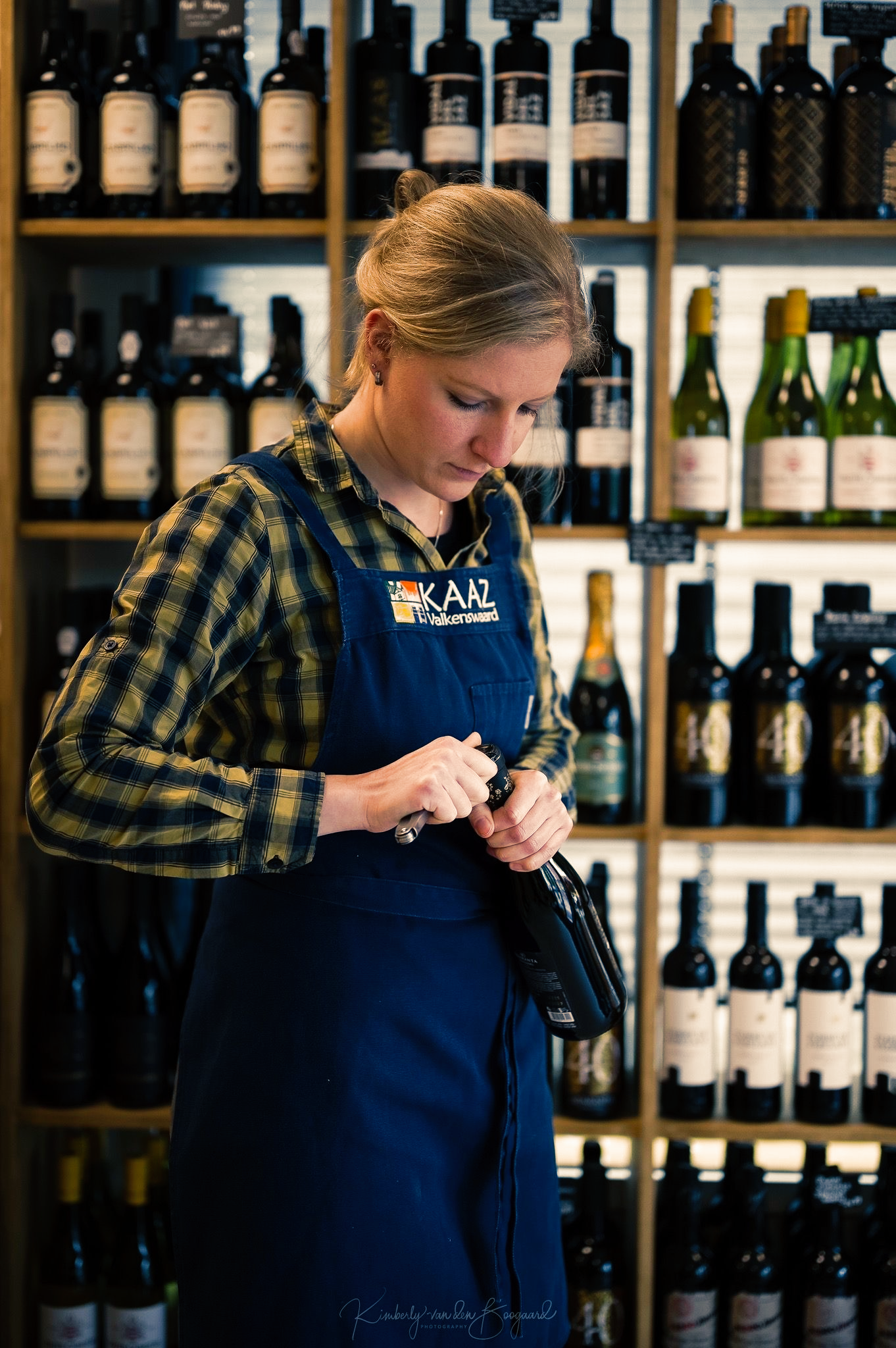 Vrouw met schort opent een wijnfles in een winkel, omringd door planken vol wijnflessen.
