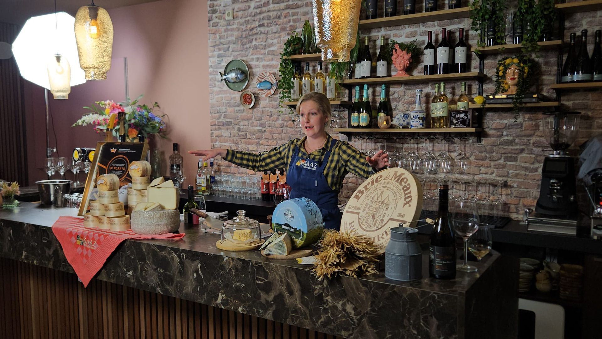 Vrouw met uitgestrekte armen aan een bar, omringd door eten en drinken, tegen een bakstenen muur op de achtergrond.