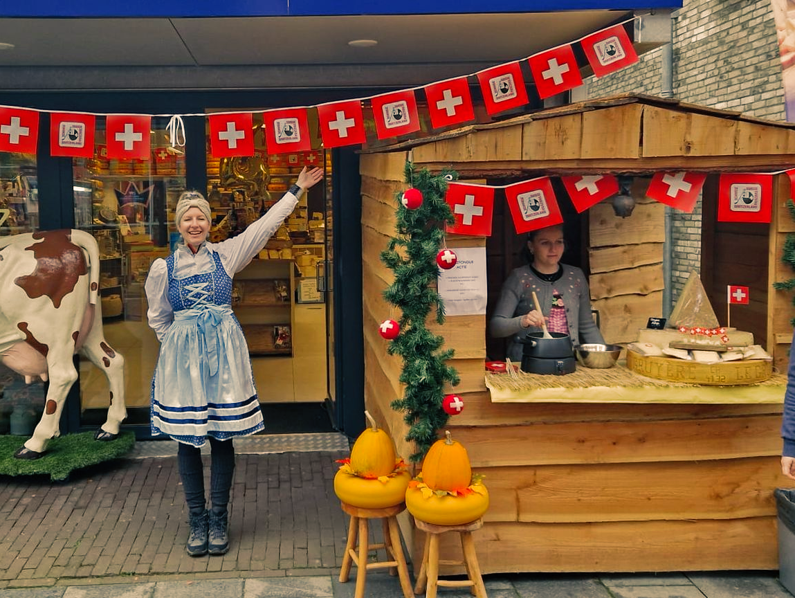 Een vrouw in Zwitserse kledij gebaart naar een houten standaard met Zwitserse vlaggen. Persoon achter de toonbank.
