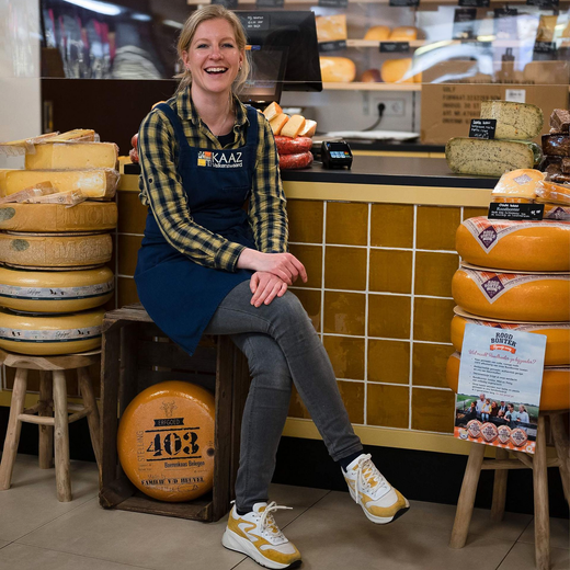 Lachende vrouw zit in een kaaswinkel, draagt een schort en is omringd door kazen.