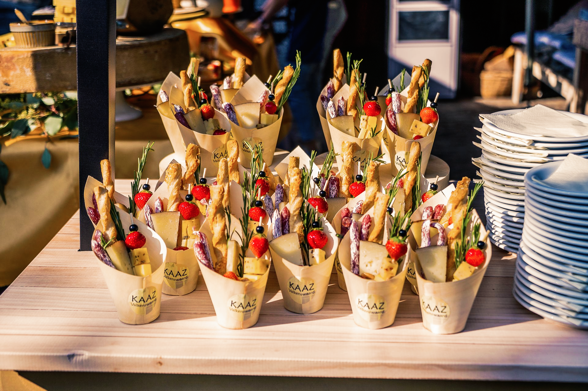 Een arrangement van hapjes in papieren puntzakken op een houten tafel. Bevat stokbrood, tomaten en paars fruit.