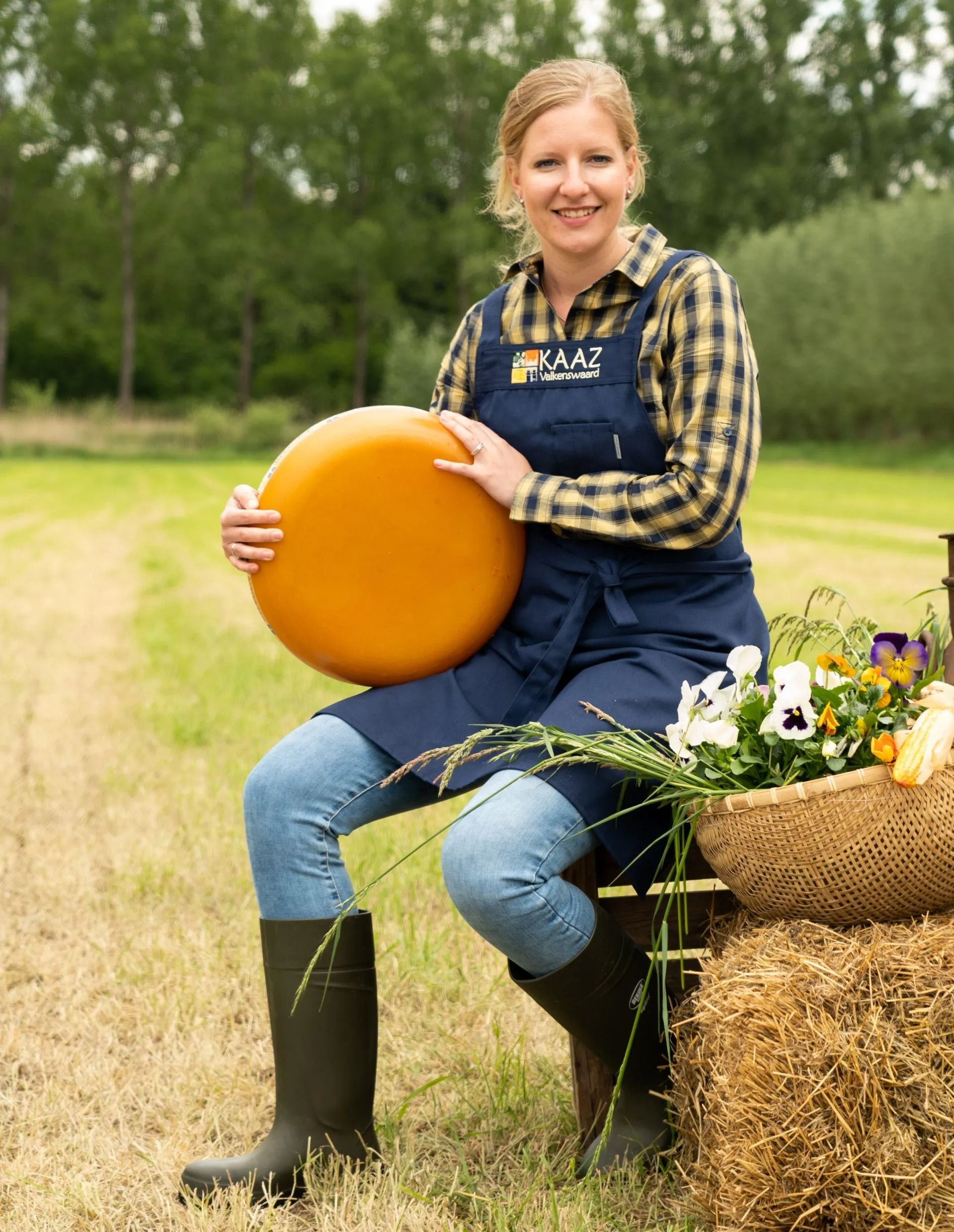 Vrouw in werkkleding met grote ronde kaas in haar handen, zittend op een hooibaal, buiten.