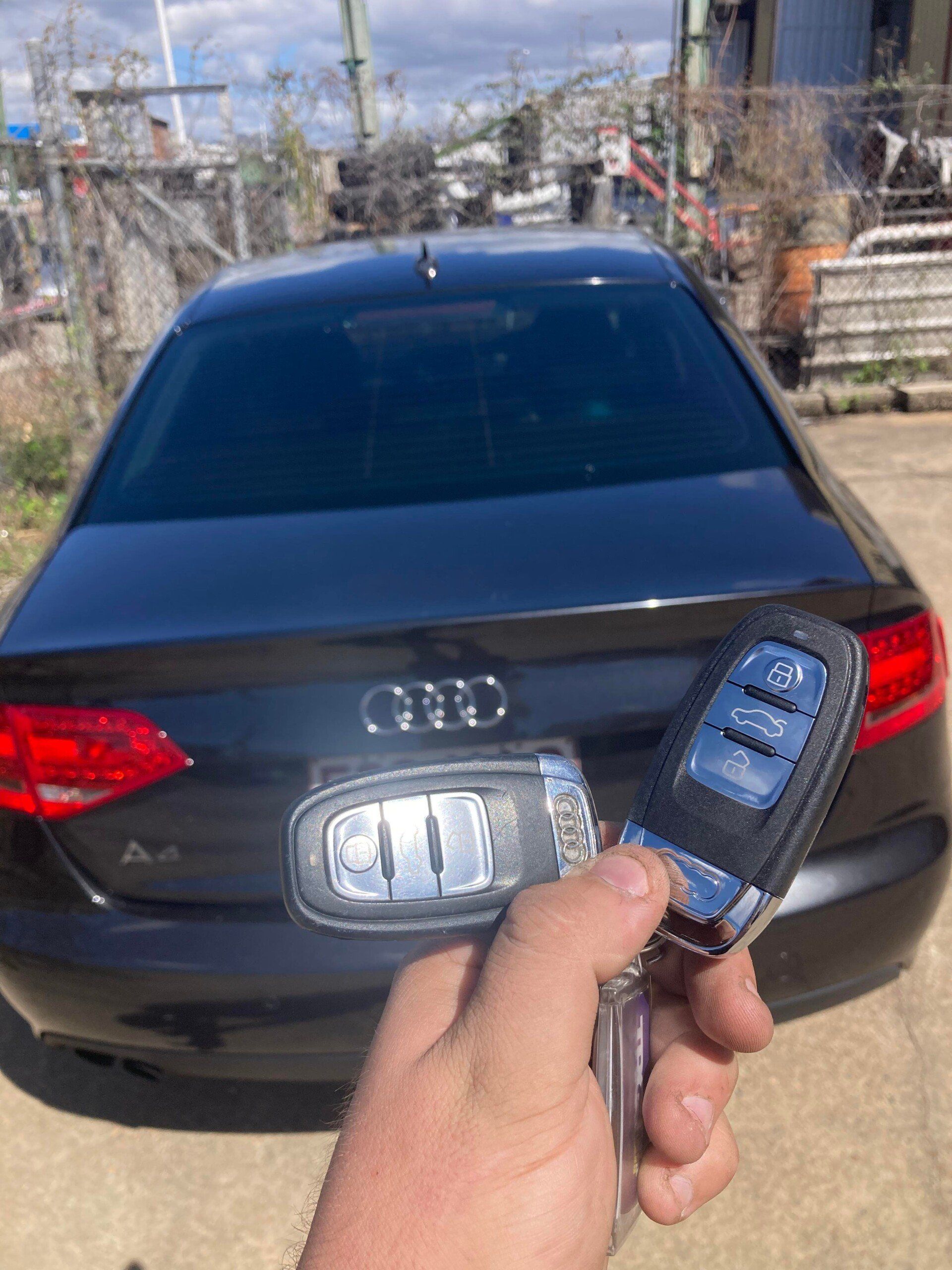 A hand holding two car keys in front of a black Audi A4 sedan, outdoors.— Locksmiths In Thuringowa Central, QLD