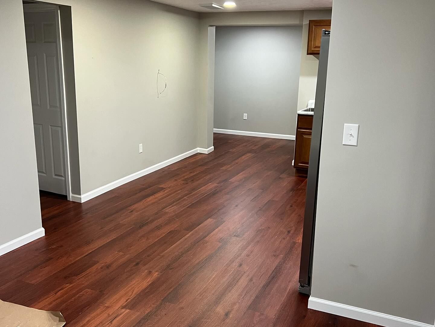 Room with dark wood flooring, gray walls, doorway on left, and kitchen cabinets visible at the end.