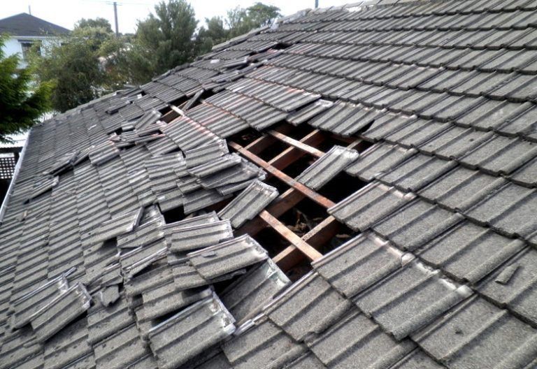 Damaged tiled roof with large hole exposing wooden structure. Gray tiles are broken and scattered.