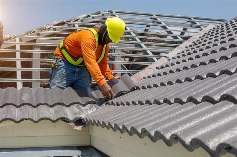 Roofer in orange shirt and yellow hard hat installing gray roof tiles on a house.