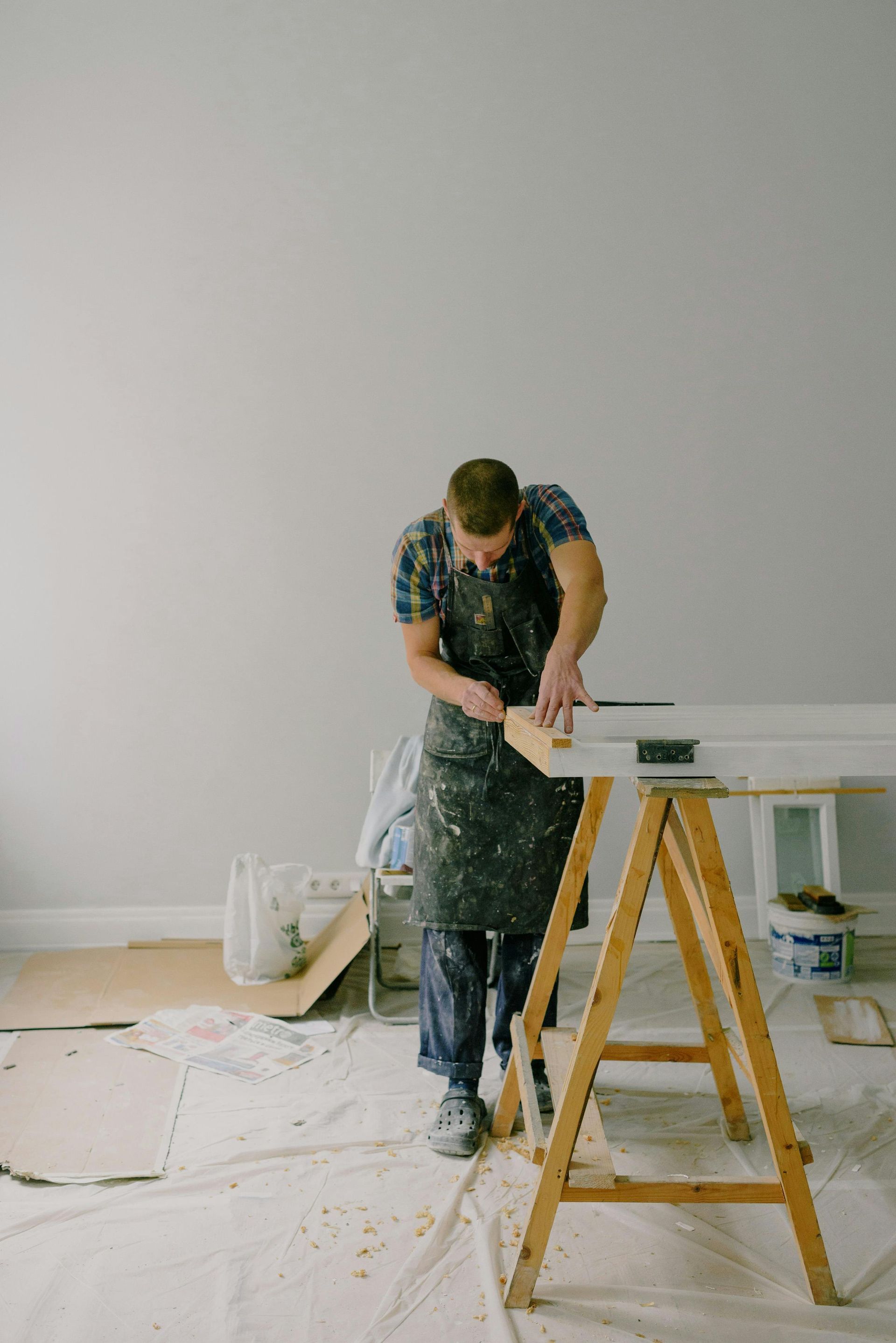 Person sanding a white board in a room under renovation, with a wooden sawhorse in the foreground