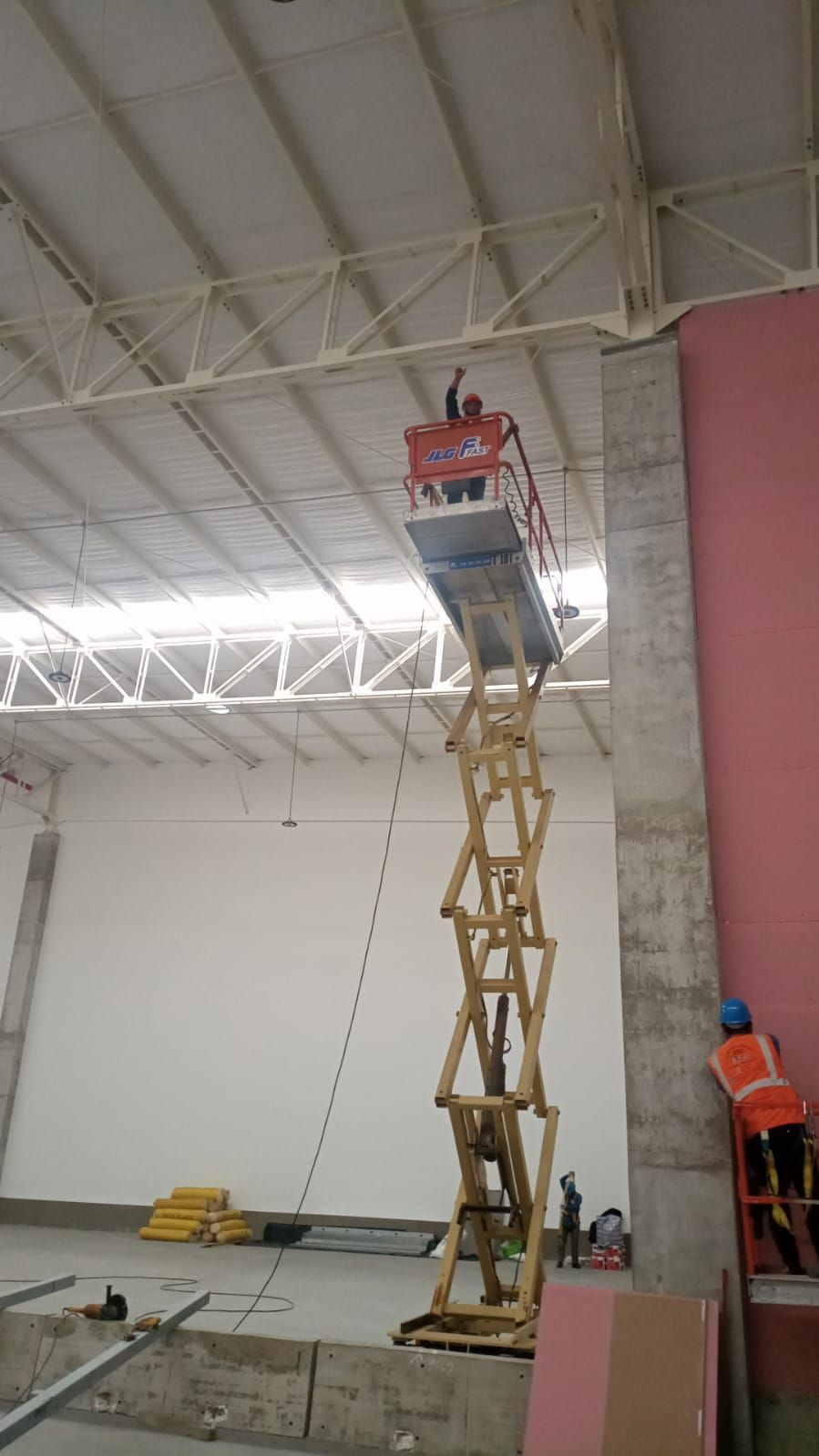 Un trabajador en una plataforma elevadora de tijera alta cerca de una pared rosa dentro de una gran habitación que parece un almacén.