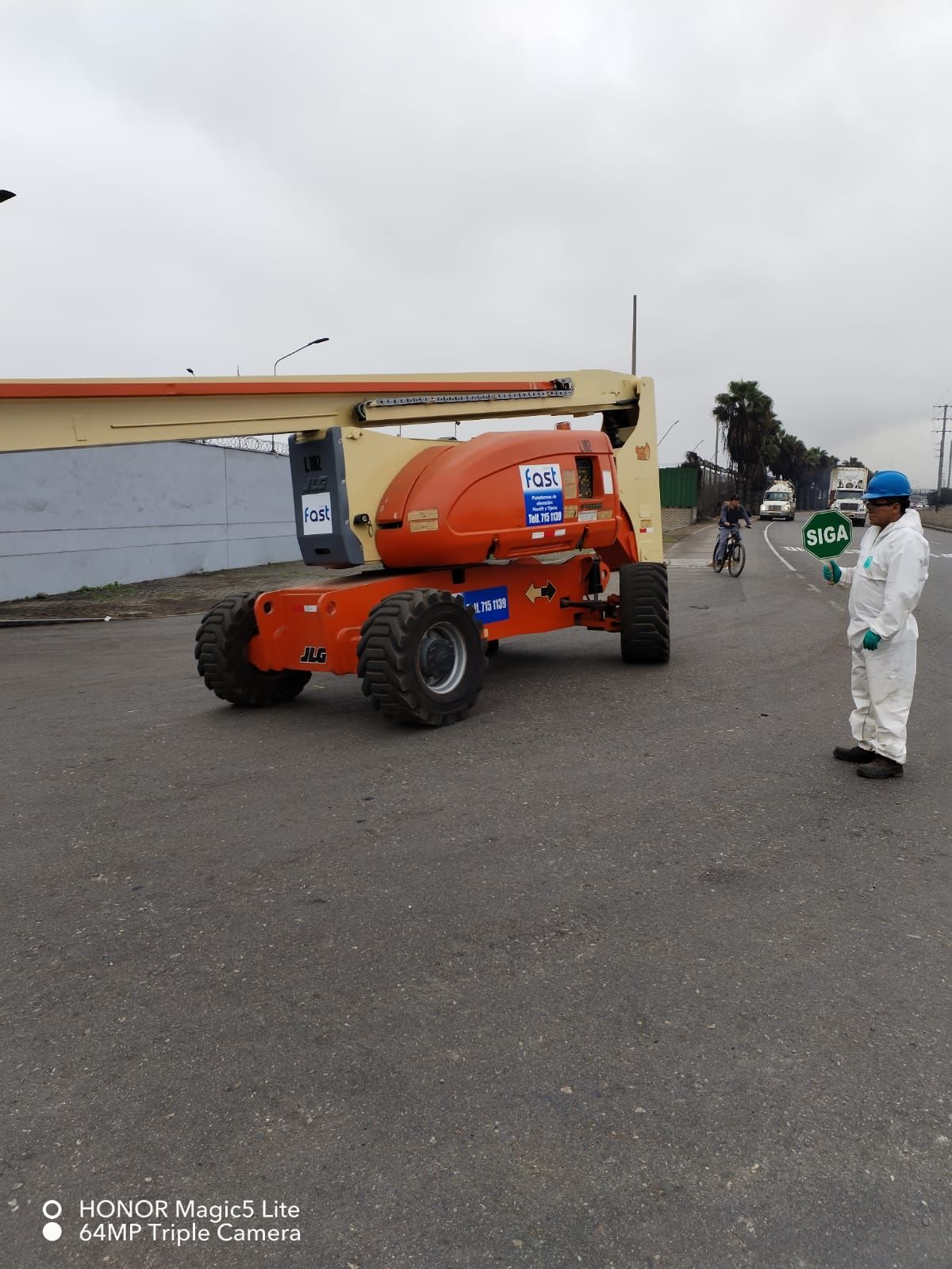 Plataforma elevadora naranja en un terreno de grava con dos trabajadores cerca bajo un cielo nublado.