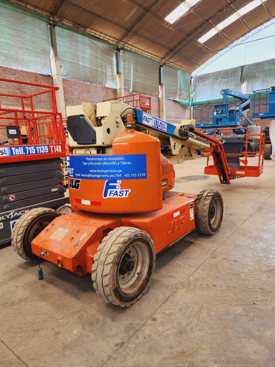 Una plataforma elevadora de brazo naranja y azul en un almacén, con otras plataformas elevadoras al fondo.