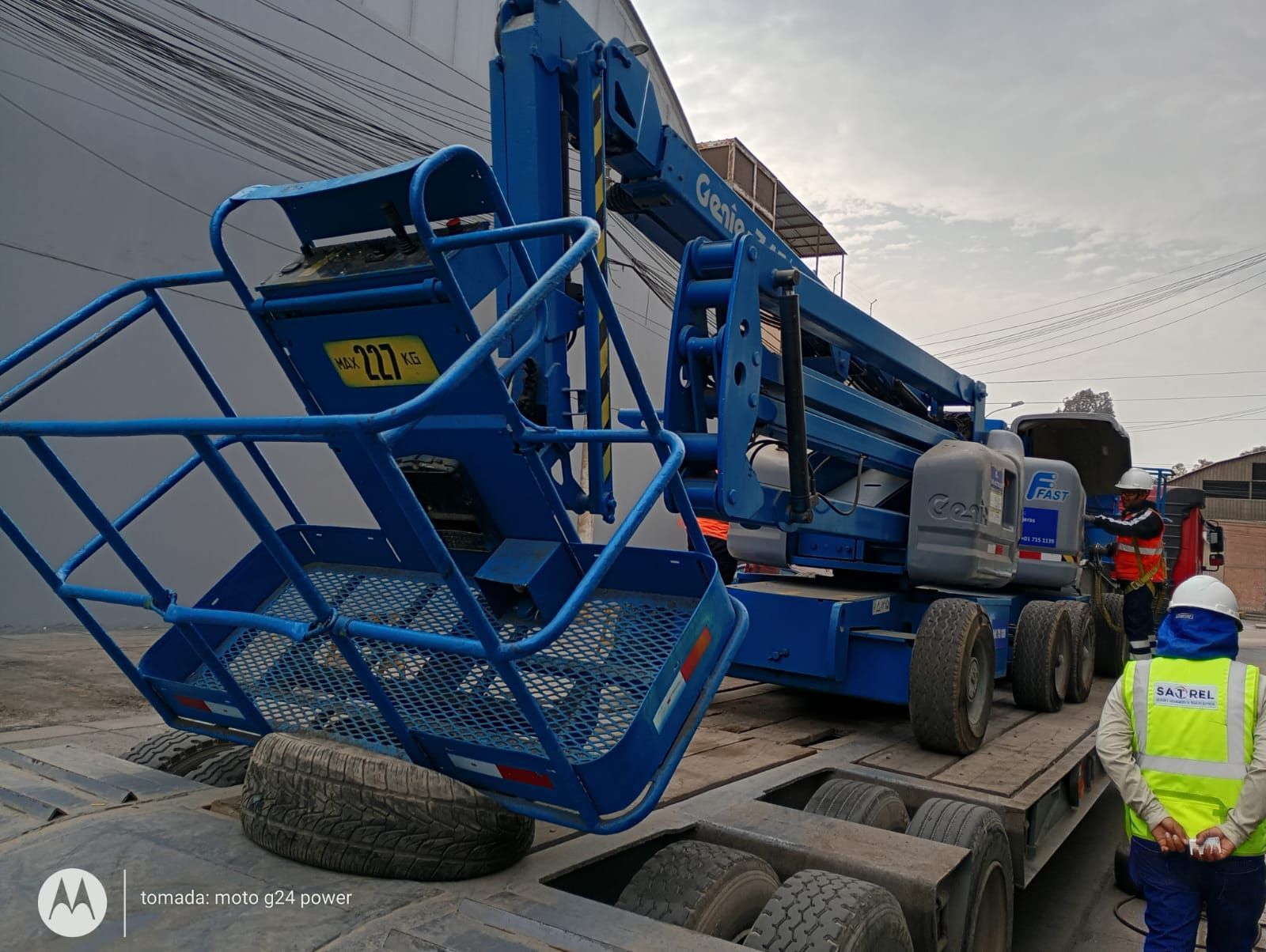 Una plataforma elevadora azul cargada en un camión cerca de un trabajador con un chaleco reflectante en la cuneta.