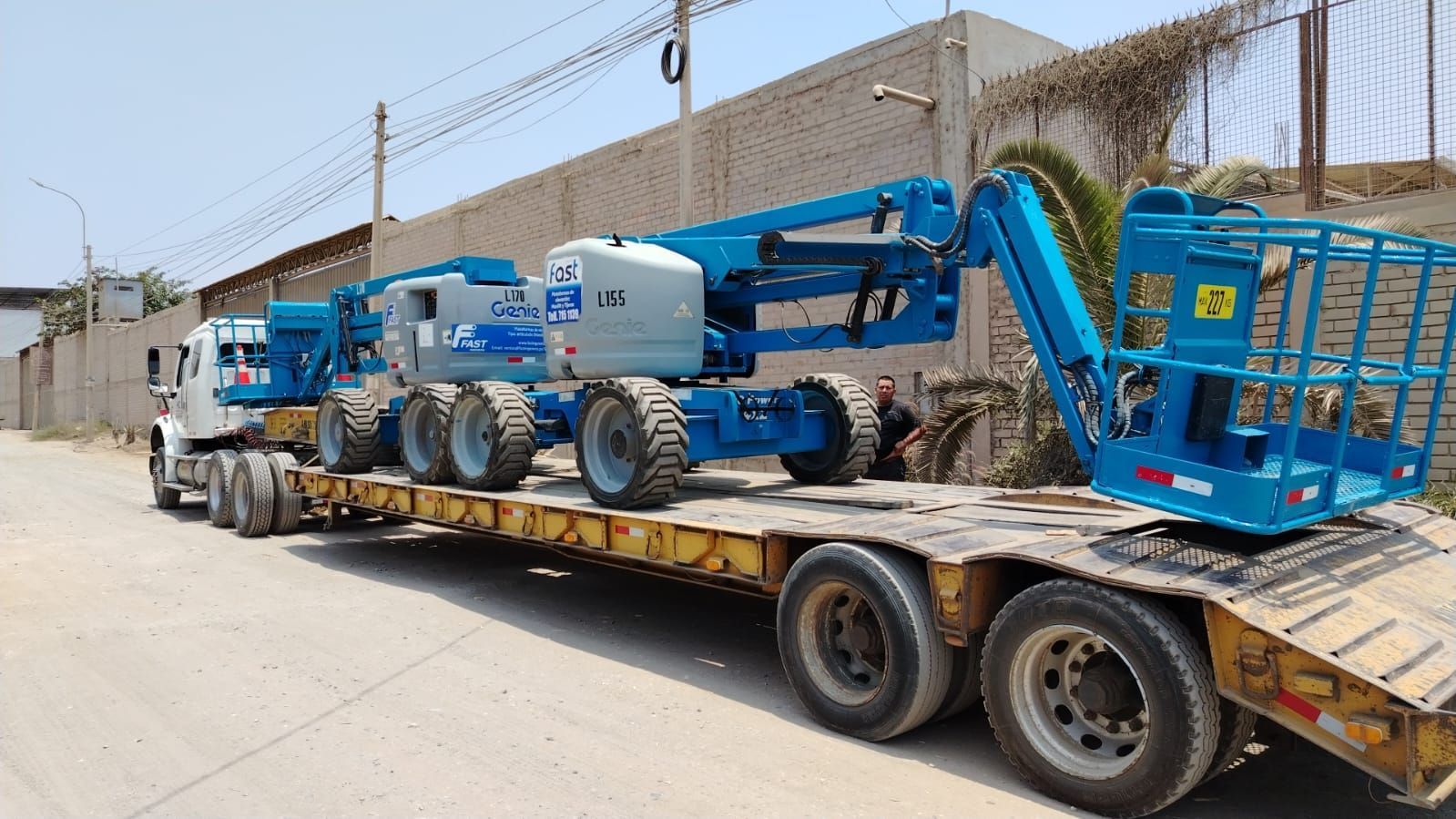 Plataforma elevadora azul montada sobre un remolque de plataforma estacionada junto a un edificio en construcción.