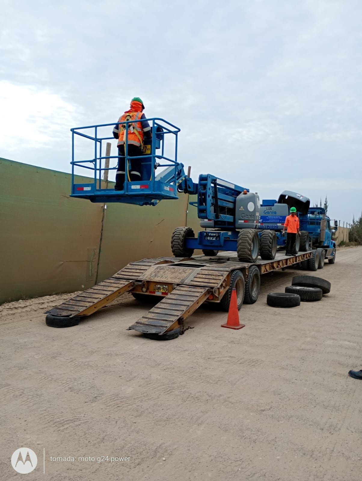 Trabajadores en una plataforma elevadora azul descargan equipo pesado en un terreno baldío mediante rampas metálicas.