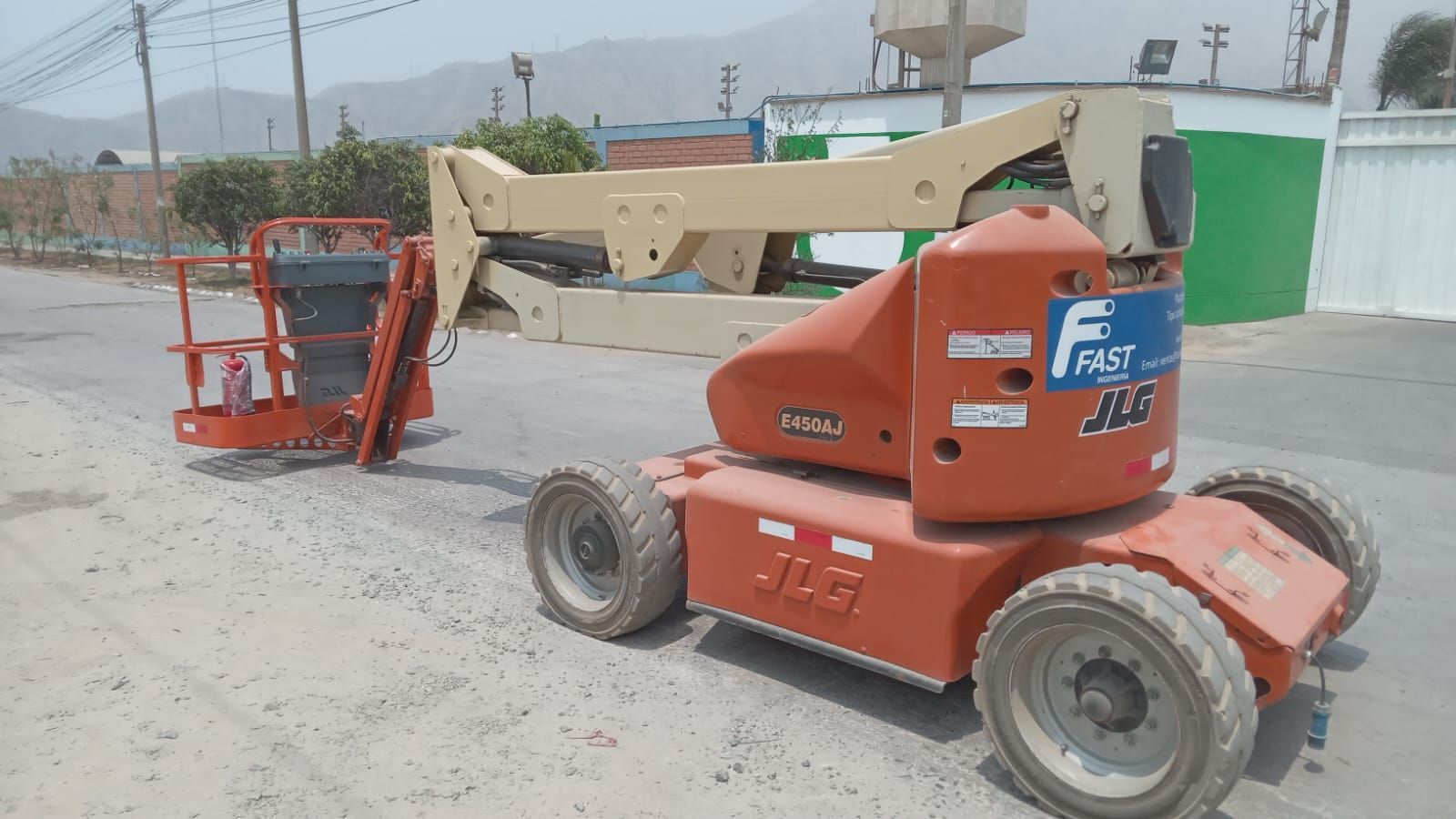 Una grúa elevadora roja y beige estacionada en un terreno polvoriento cerca de un edificio verde.
