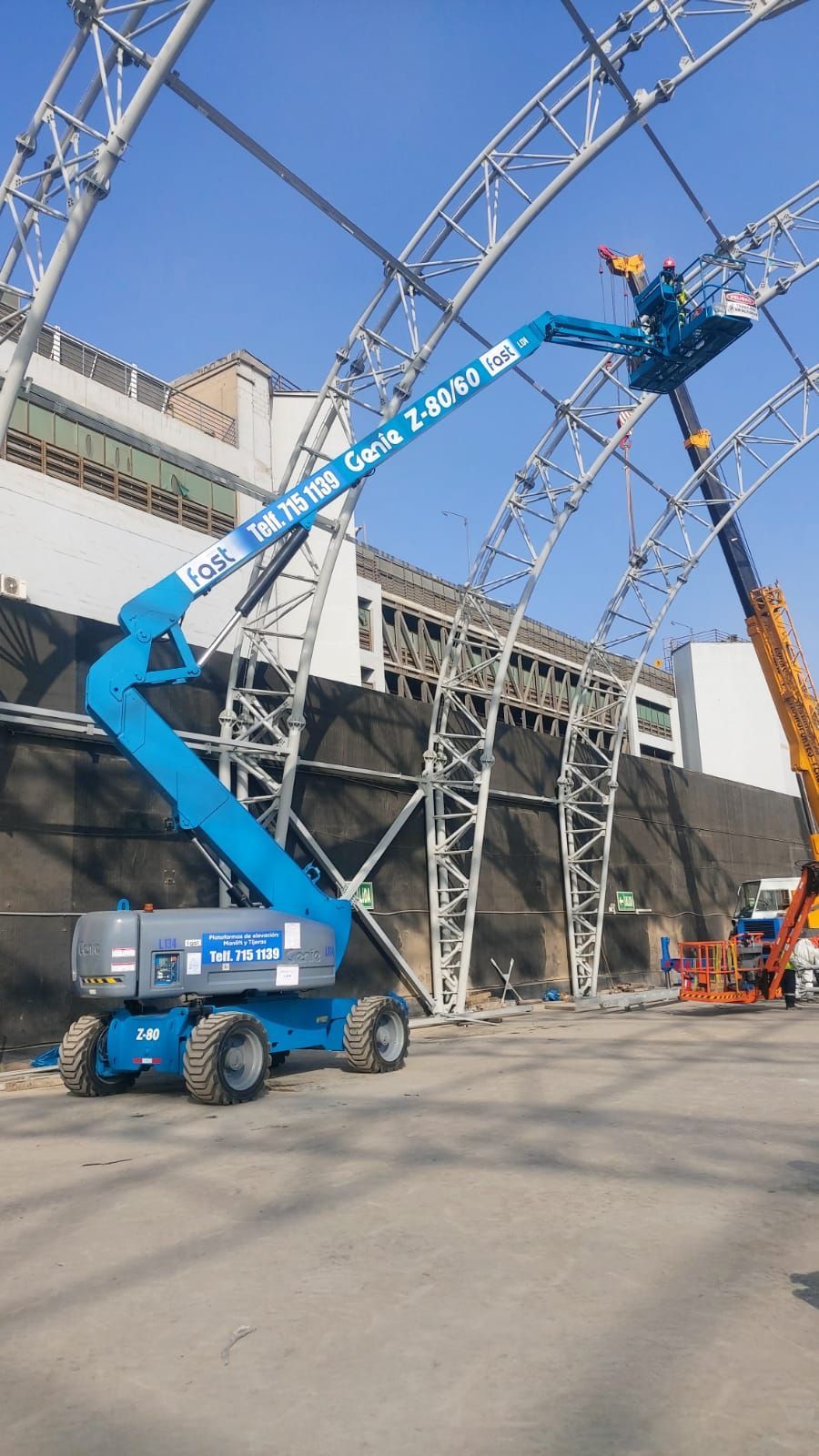Una plataforma elevadora azul se eleva hasta los trabajadores que se encuentran en una estructura metálica junto a un edificio.