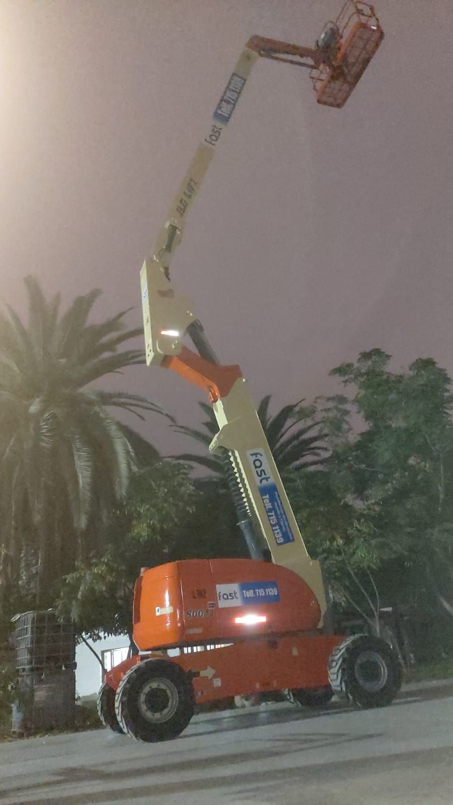 Una grúa elevadora naranja se extiende a gran altura junto a las palmeras al anochecer o por la noche.