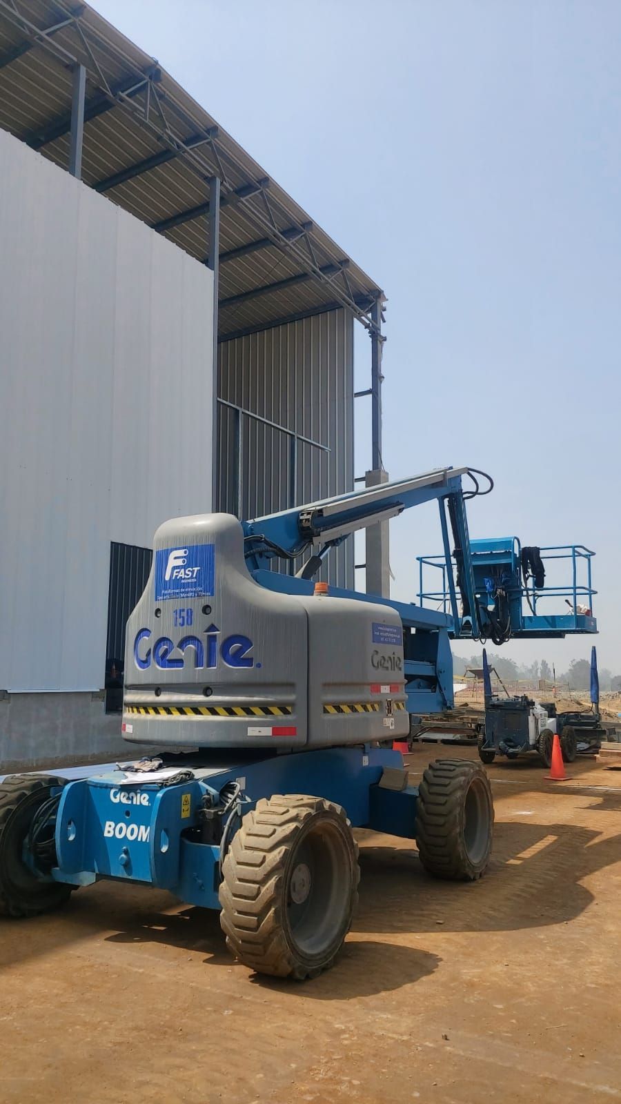 Una plataforma elevadora telescópica Blue Genie está estacionada junto a un gran almacén metálico en una obra de construcción de tierra.
