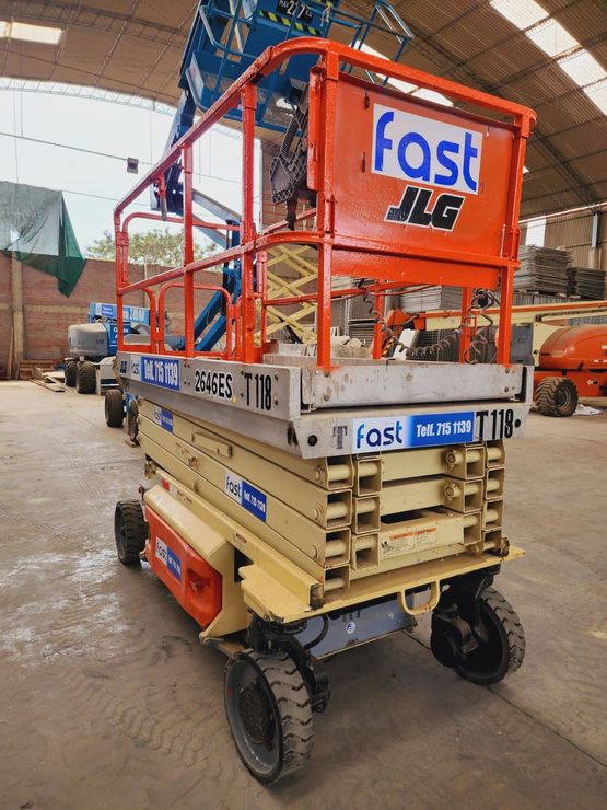 Obrero de la construcción en una plataforma elevadora de tijera naranja en una obra, con la excavadora y el equipo de seguridad a la vista.