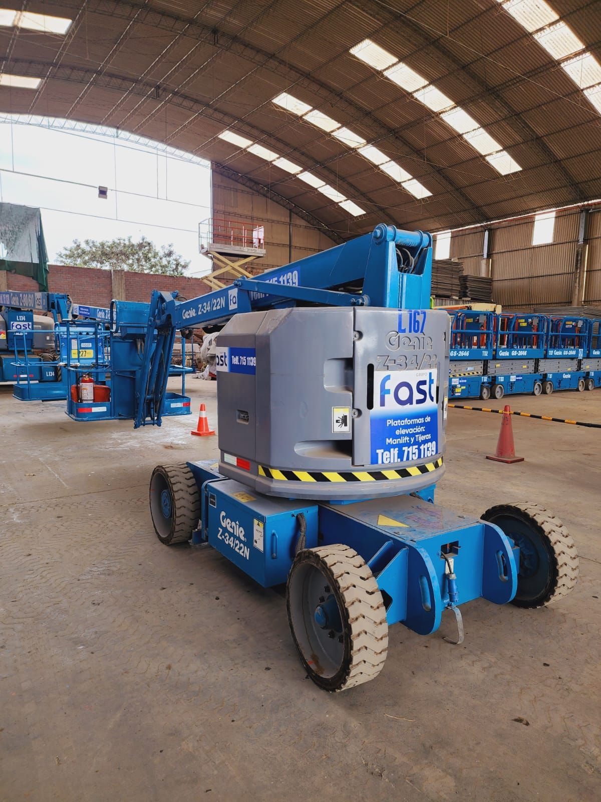 Plataforma elevadora móvil azul y gris en un almacén interior con equipos apilados al fondo.
