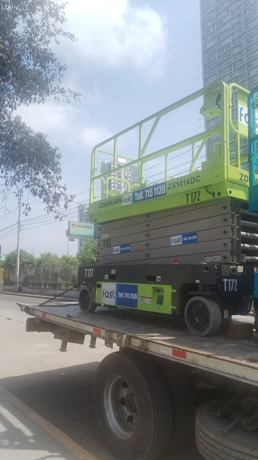 Plataforma elevadora de tijera verde sobre un camión de plataforma en una calle de la ciudad junto a edificios altos y árboles.