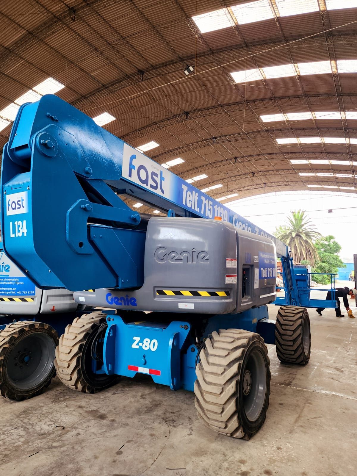 Plataforma elevadora azul con brazos articulados, ruedas grandes y brazo elevado, ubicada dentro de un almacén.