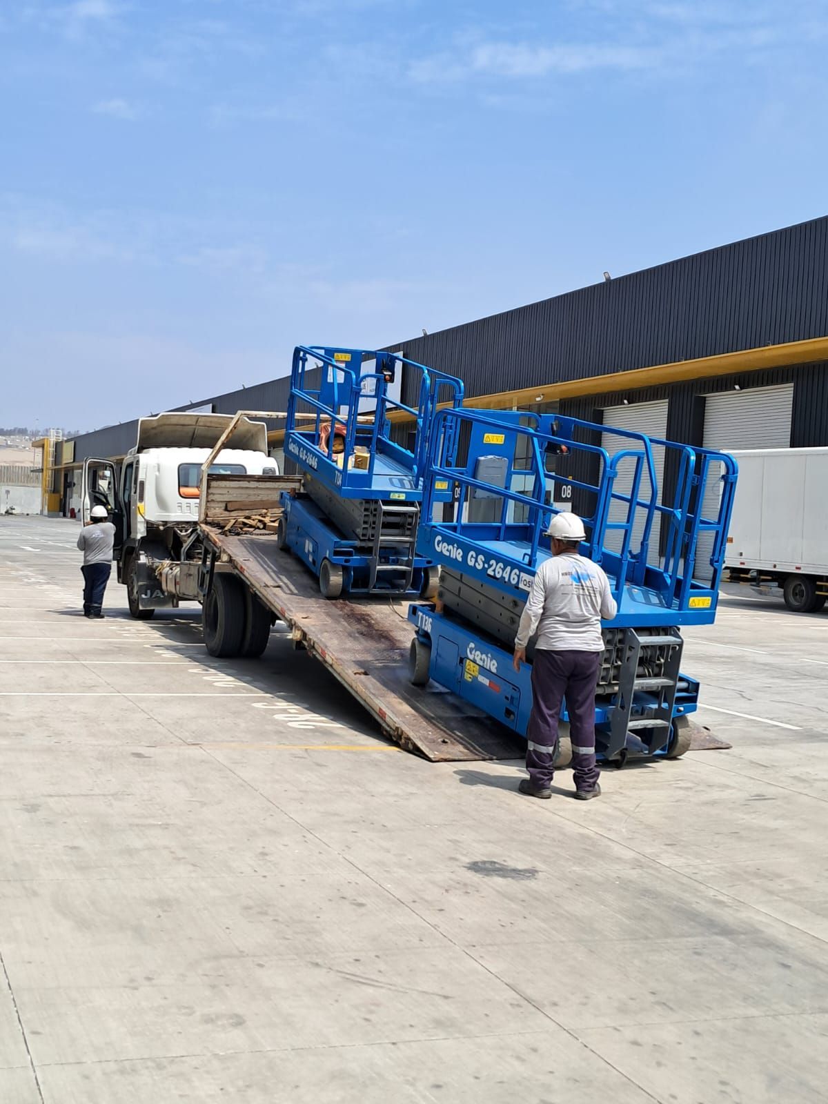 Unas escaleras azules del aeropuerto están siendo cargadas en un camión de plataforma, con trabajadores cerca en una pista soleada.