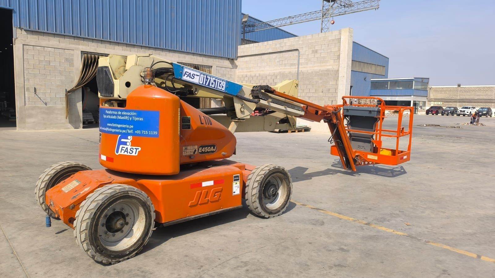 Una plataforma elevadora naranja está estacionada en un terreno de hormigón junto a edificios industriales.
