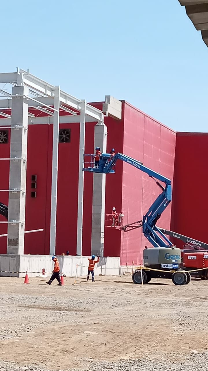 Plataforma elevadora azul junto a un edificio rojo en construcción con estructura de acero blanco a la vista.